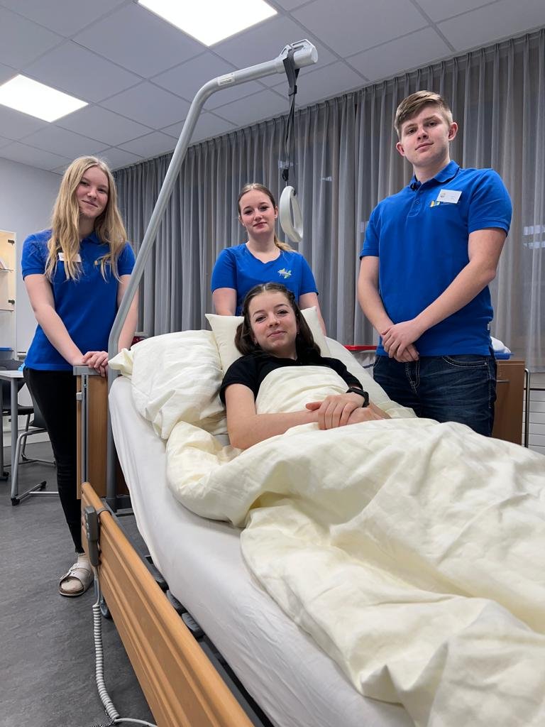 Drei SchülerInnen stehen um ein Pflegebett mit einer Schülerin die eine pflegebedürftige Person darstellt.